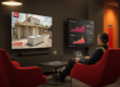 Man sitting on sofa watching sky, another tv next to it displaying graphs